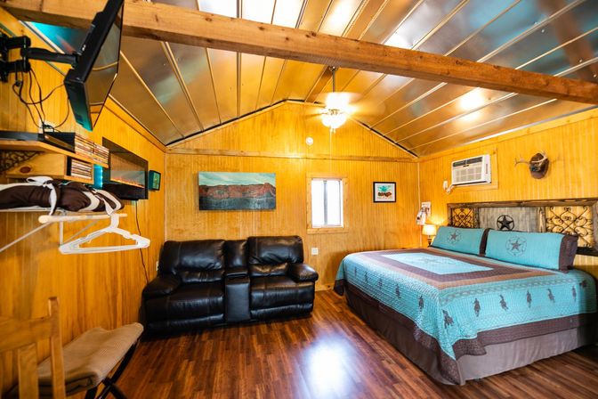Cozy rustic cabin bedroom with wood-paneled walls and metal vaulted ceiling, turquoise king bed, black leather loveseat, hardwood floors, wall air conditioner