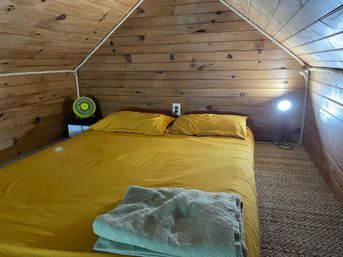 Cozy cabin-style attic loft with sloped wood-paneled walls, low bed dressed in mustard yellow sheets and pillows, folded blanket, small green fan and bedside lamp on a carpeted floor.
