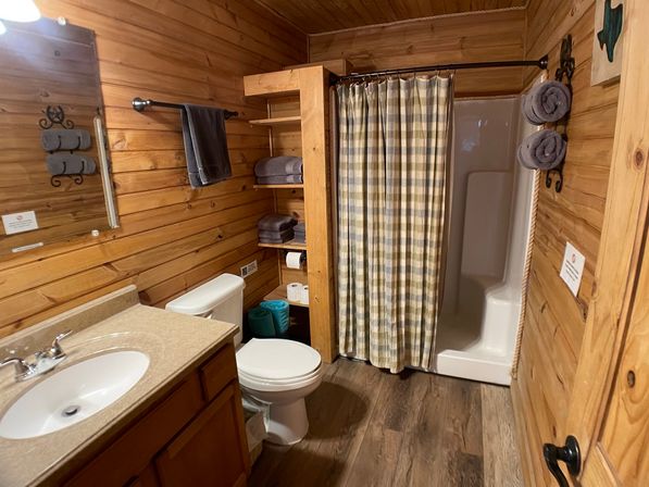 Cozy rustic wood-paneled cabin bathroom — vanity sink, toilet, shower with plaid curtain, open shelves stocked with folded towels and toilet paper