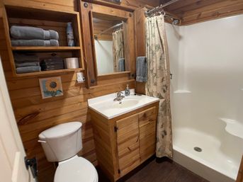 Cozy rustic cabin bathroom with wood-paneled walls, white toilet, wood vanity and mirror, stacked gray towels on open shelves, sunflower wall art, and a shower with beige nature-print curtain.
