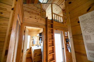 Cozy tiny cabin interior with warm pine paneling, a wooden ladder leading to a loft, a glass entry door, and a compact bathroom vanity with towels and shelves.