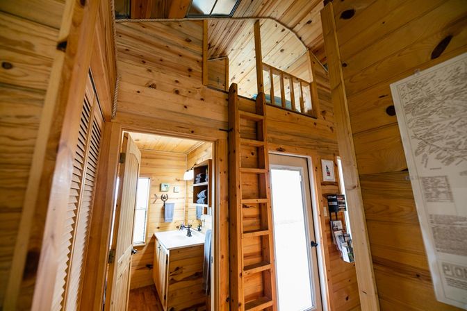 Cozy tiny cabin interior with warm pine paneling, a wooden ladder leading to a loft, a glass entry door, and a compact bathroom vanity with towels and shelves.