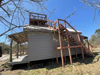 Raised countryside cabin with pale siding, covered porch and wooden stairway up to a rooftop deck, surrounded by budding trees under a clear blue sky.