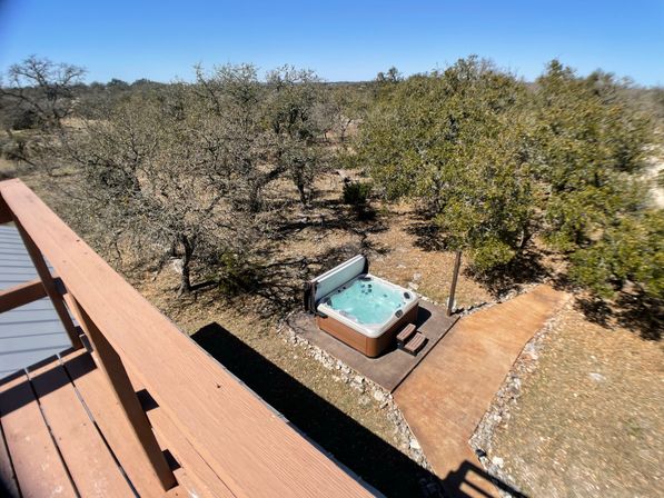 Balcony view of a bubbling outdoor hot tub on a concrete pad beside a curved, stone-lined path, set in a sunlit, oak‑scattered rural backyard.