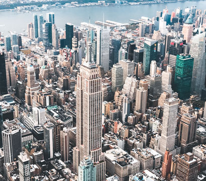 Aerial view of New York City skyline — Empire State Building rising among dense Manhattan skyscrapers with the Hudson River waterfront in the background.