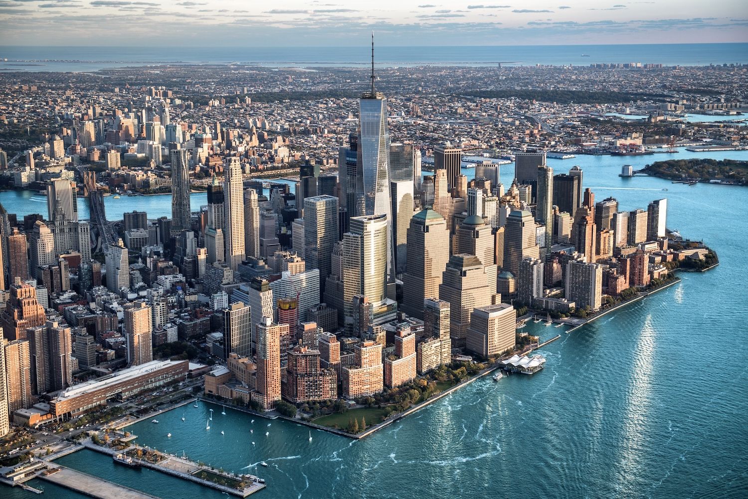 Aerial view of Lower Manhattan, New York City skyline with One World Trade Center towering above dense skyscrapers, waterfront piers and boats on blue harbor waters.