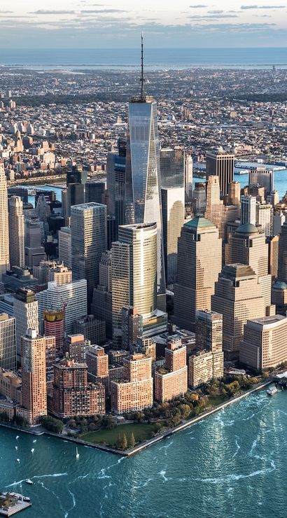 Aerial view of Lower Manhattan, New York City skyline with One World Trade Center towering above dense skyscrapers, waterfront piers and boats on blue harbor waters.