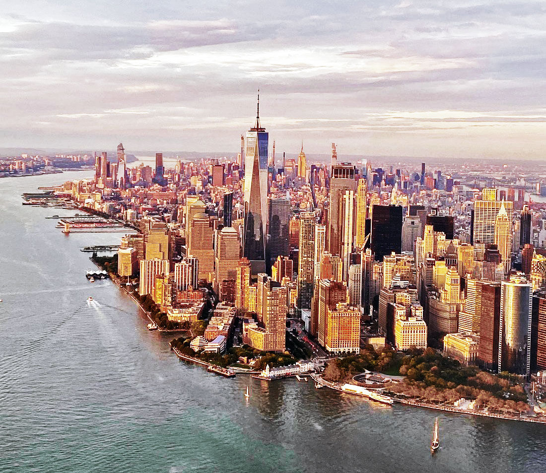 Aerial view of Lower Manhattan, New York City at golden hour with One World Trade Center rising above skyscrapers, Hudson River shoreline and small boats.