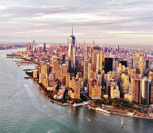 Aerial view of Lower Manhattan, New York City at golden hour with One World Trade Center rising above skyscrapers, Hudson River shoreline and small boats.