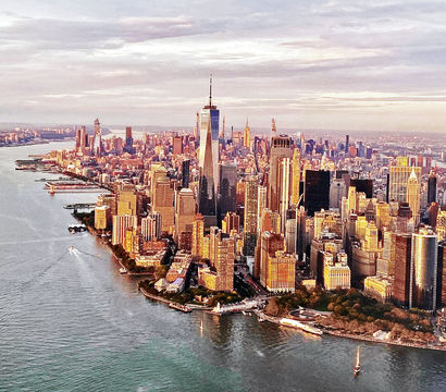 Aerial view of Lower Manhattan, New York City at golden hour with One World Trade Center rising above skyscrapers, Hudson River shoreline and small boats.