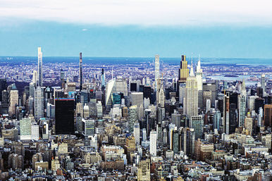 Aerial view of New York City Midtown Manhattan skyline with dense skyscrapers, distant rivers and a bright blue sky.