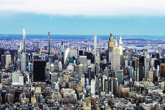 Aerial view of New York City Midtown Manhattan skyline with dense skyscrapers, distant rivers and a bright blue sky.