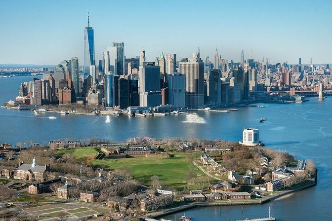 Aerial view of Governors Island green lawns in the foreground with Lower Manhattan skyline and One World Trade Center rising over New York Harbor on a clear blue day, ferries crossing the water