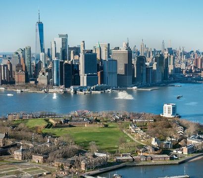 Aerial view of Governors Island green lawns in the foreground with Lower Manhattan skyline and One World Trade Center rising over New York Harbor on a clear blue day, ferries crossing the water