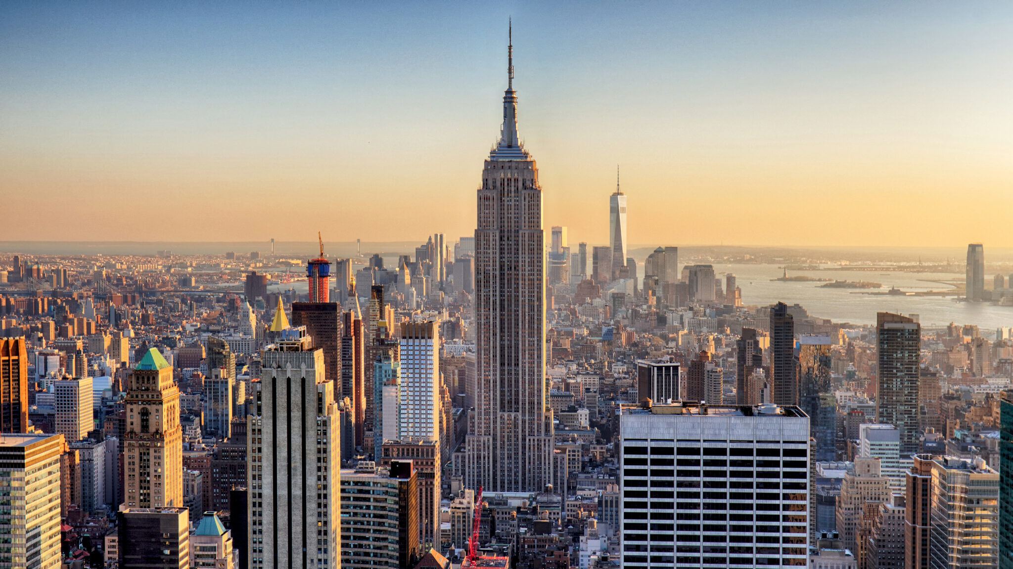 Aerial view of New York City skyline at golden sunset, Empire State Building centered with One World Trade Center and the Statue of Liberty visible in the distance.