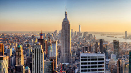 Aerial view of New York City skyline at golden sunset, Empire State Building centered with One World Trade Center and the Statue of Liberty visible in the distance.