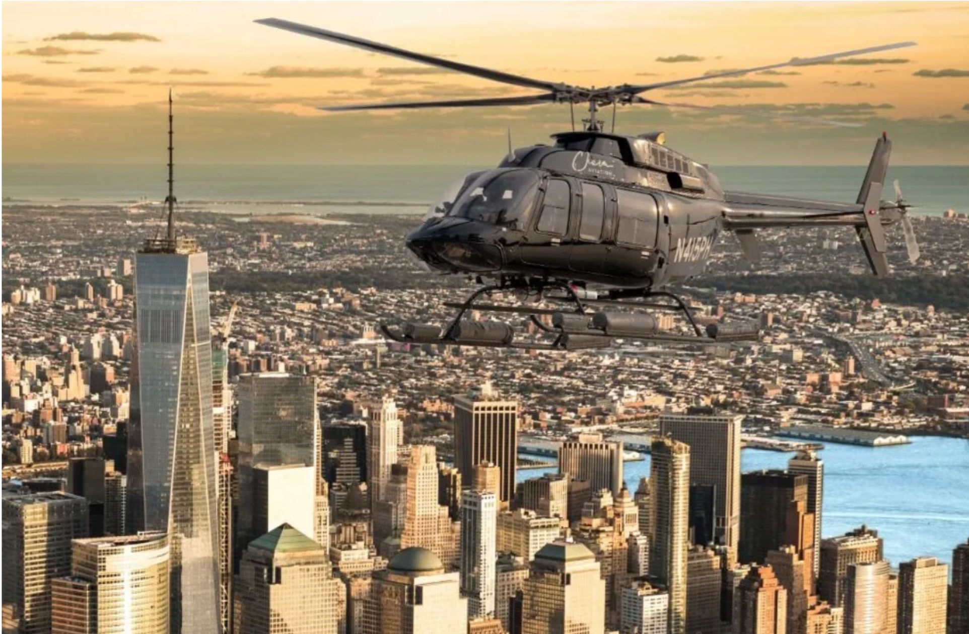Black helicopter flying over Lower Manhattan skyline at golden sunset, with skyscrapers, waterfront and distant coastline visible beneath.