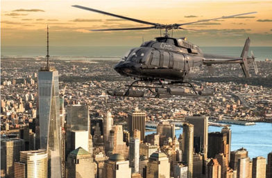 Black helicopter flying over Lower Manhattan skyline at golden sunset, with skyscrapers, waterfront and distant coastline visible beneath.