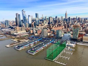 Aerial view of Manhattan skyline along the Hudson River, colorful waterfront piers and marinas in the foreground with dense skyscrapers and a bright blue sky.