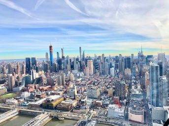 Aerial view of Midtown Manhattan skyline with dense high-rise skyscrapers, waterfront piers and a wide blue sky streaked with clouds.
