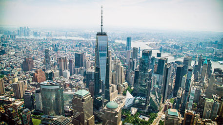 Aerial, sky-high view of Lower Manhattan, New York City skyline featuring a prominent glass skyscraper, dense city blocks, rivers and bridges.