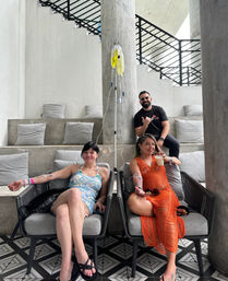 Three adults relaxing in a modern resort-style wellness lounge — two women seated in chairs receiving IV drip therapy while one sips a drink and a man poses behind; concrete pillars, staircase railings and geometric tile floor visible.