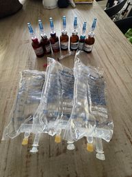 Amber vials with blue infusion caps and a syringe lined up behind four clear IV fluid bags on a rustic wooden table