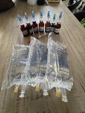 Amber vials with blue infusion caps and a syringe lined up behind four clear IV fluid bags on a rustic wooden table
