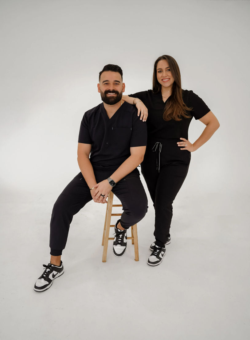 Friendly healthcare team portrait — two clinicians in black scrubs, one seated on a wooden stool and one standing with a hand on their shoulder against a white studio background.