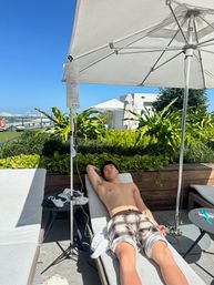 Shirtless person relaxing on a rooftop pool lounge chair under a white umbrella with an IV drip bag nearby, tropical plants and urban skyline under a bright blue sky.