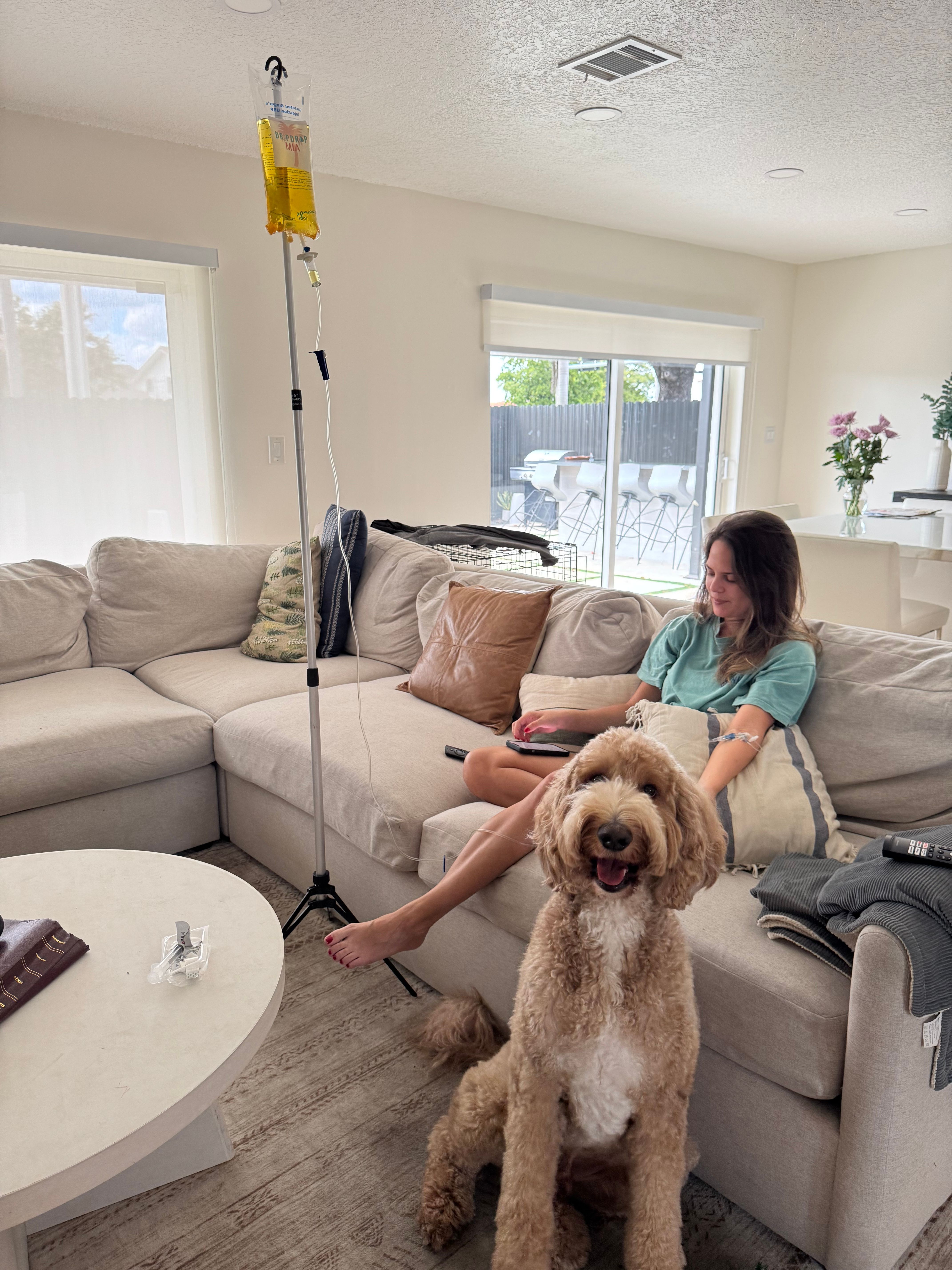 Smiling goldendoodle in the foreground while a woman relaxes on a beige sectional receiving IV therapy from a yellow IV bag on a pole in a bright, sunlit living room with sliding glass doors.