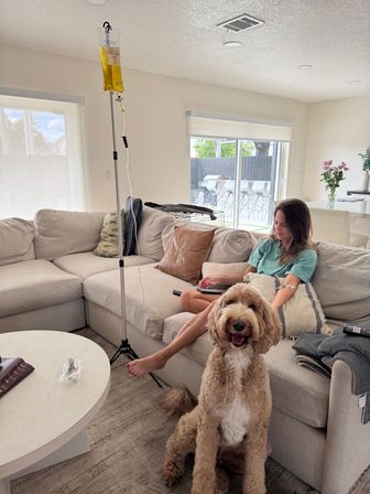 Smiling goldendoodle in the foreground while a woman relaxes on a beige sectional receiving IV therapy from a yellow IV bag on a pole in a bright, sunlit living room with sliding glass doors.