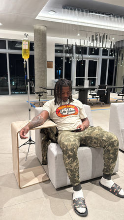 Person receiving IV drip in a modern high-rise lounge at night, seated on a gray armchair wearing a graphic tee, camo pants and slide sandals with socks, looking at a phone; yellow IV bag on a stand and contemporary pendant lights overhead.