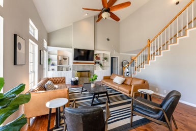 Sunlit open living room with high vaulted ceiling and wooden ceiling fan, cognac leather sofas and black armchairs around a striped rug and coffee table, built-in fireplace and white staircase — bright, modern and cozy.