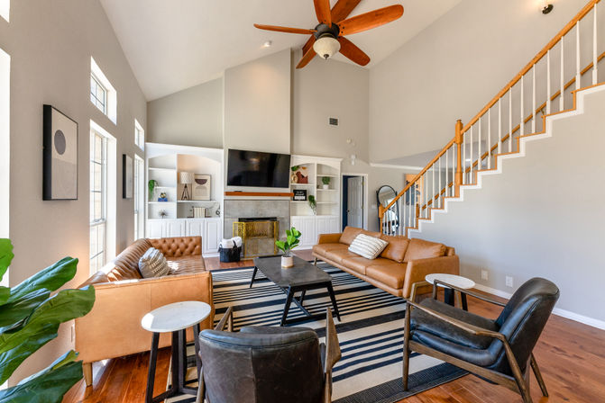 Sunlit open living room with high vaulted ceiling and wooden ceiling fan, cognac leather sofas and black armchairs around a striped rug and coffee table, built-in fireplace and white staircase — bright, modern and cozy.
