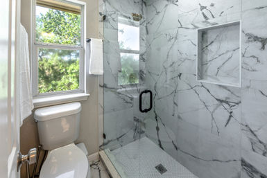 Bright residential bathroom with white toilet and modern glass-enclosed marble tile shower, hex-tile floor, recessed niche, black handle and a window with green outdoor view.