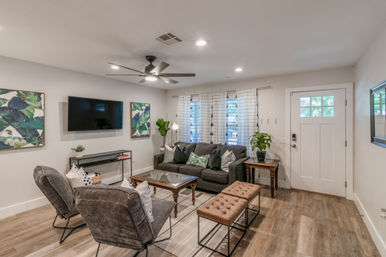 Cozy modern living room with gray sofa, two accent chairs, wall-mounted TV, ceiling fan, tufted bench, wood floors and white front door