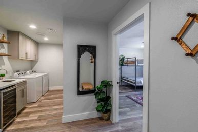 Bright modern home interior: laundry area with side-by-side washer and dryer, gray cabinets and wine fridge, hallway with decorative mirror and potted plant leading to a bunk-bed bedroom on wood-look floors.