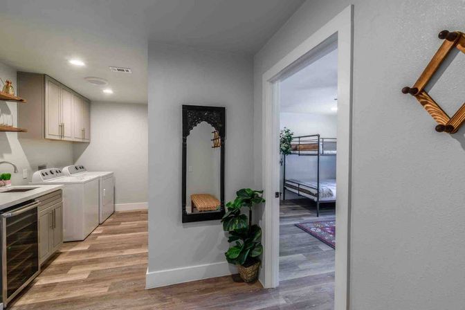 Bright modern home interior: laundry area with side-by-side washer and dryer, gray cabinets and wine fridge, hallway with decorative mirror and potted plant leading to a bunk-bed bedroom on wood-look floors.