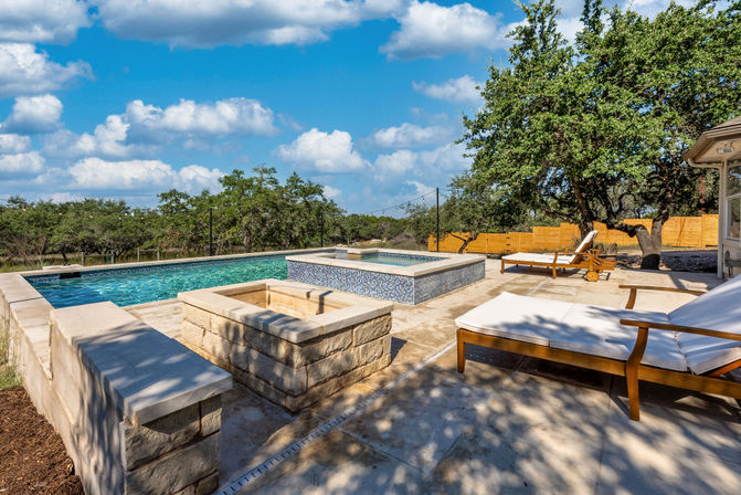 Sunny backyard pool with raised mosaic-tiled spa, stone fire pit and wooden lounge chairs on a travertine patio beneath live oak trees and a blue sky