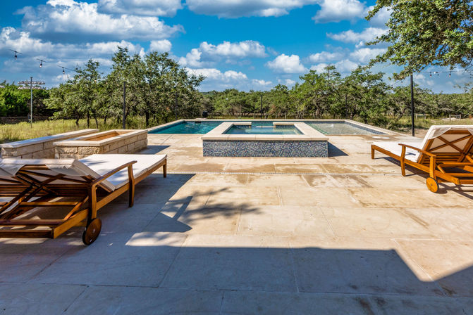 Sunlit stone patio with resort-style rectangular pool and raised spa, wooden chaise lounges on wheels, string lights, and a treed countryside under a bright blue sky.