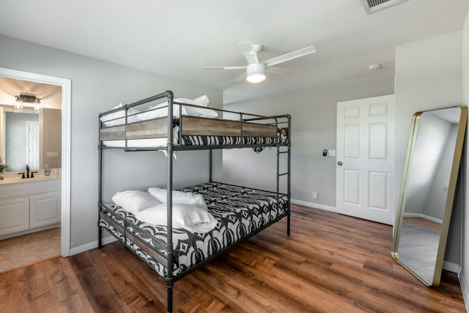 Bright modern guest bedroom with black metal bunk bed (double bottom, twin top) dressed in black-and-white patterned bedding, wood-look floors, neutral gray walls, ceiling fan, leaning full-length mirror and open doorway to an en-suite bathroom.
