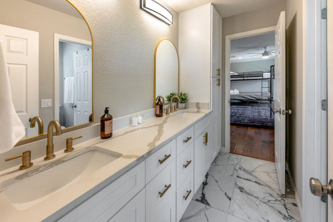 Bright modern residential bathroom with a white double vanity, quartz countertop, brass faucets, arched gold-framed mirrors, marble-look floor tile, and an open door leading to a bedroom with bunk beds.