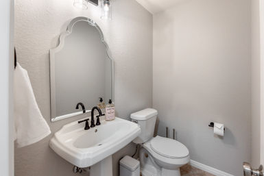 Bright modern powder room with a white pedestal sink, arched decorative mirror, matte black faucet and fixtures, white toilet, hand towel and liquid soap on neutral gray walls and tiled floor — compact guest bathroom.