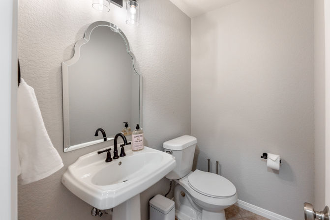 Bright modern powder room with a white pedestal sink, arched decorative mirror, matte black faucet and fixtures, white toilet, hand towel and liquid soap on neutral gray walls and tiled floor — compact guest bathroom.
