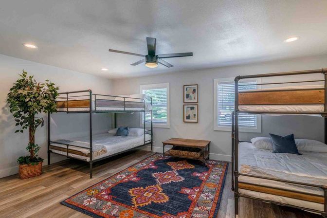 Bright modern family bunk room with two metal-frame bunk beds, colorful patterned area rug, wood floors, ceiling fan, potted plant, windows with blinds and simple wall art.