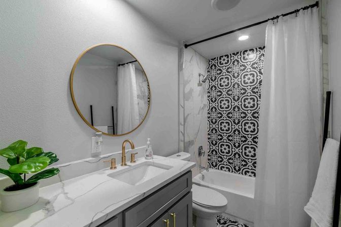 Sleek modern bathroom with marble-look countertop, round gold-framed mirror, brass faucet and gray vanity, potted green plant, and bathtub with black-and-white patterned tile and white shower curtain.