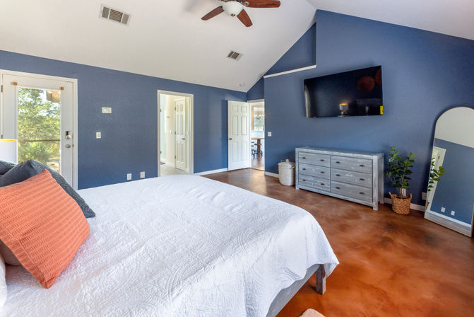 Spacious master bedroom with vaulted ceiling and navy blue walls, white quilted bed with coral accent pillow, wall-mounted TV, gray dresser, full-length mirror, potted plant, ceiling fan, and glass patio door