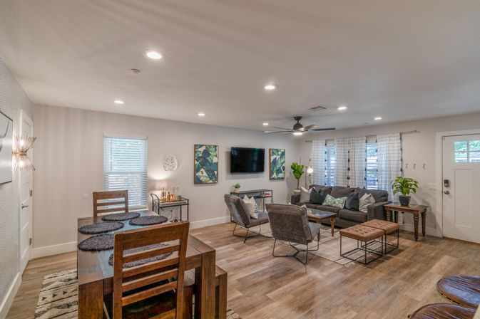 Bright, cozy open-concept living and dining room with warm wood floors, gray sectional sofa, wall-mounted TV, wooden dining table, ceiling fan, leafy artwork and potted plants.