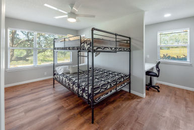 Bright, sunlit bedroom with a black metal twin-over-full bunk bed and patterned bedding, wood-look floors, large windows with blinds, white ceiling fan, and a small desk and chair in a study nook.